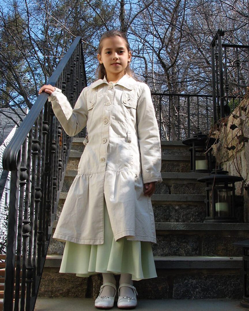 Daniella as a young girl wearing a white dress coat, standing on steps with hand on railing