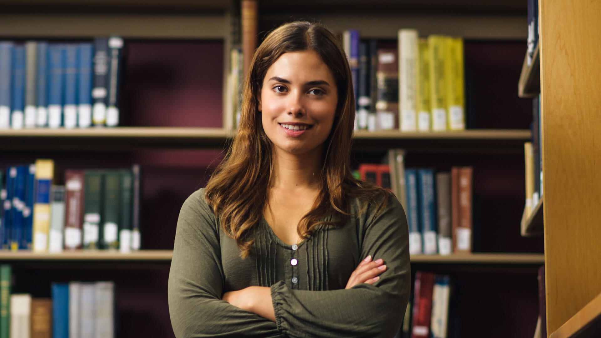 Student stands in front of book stacks at a library, arms crossed.