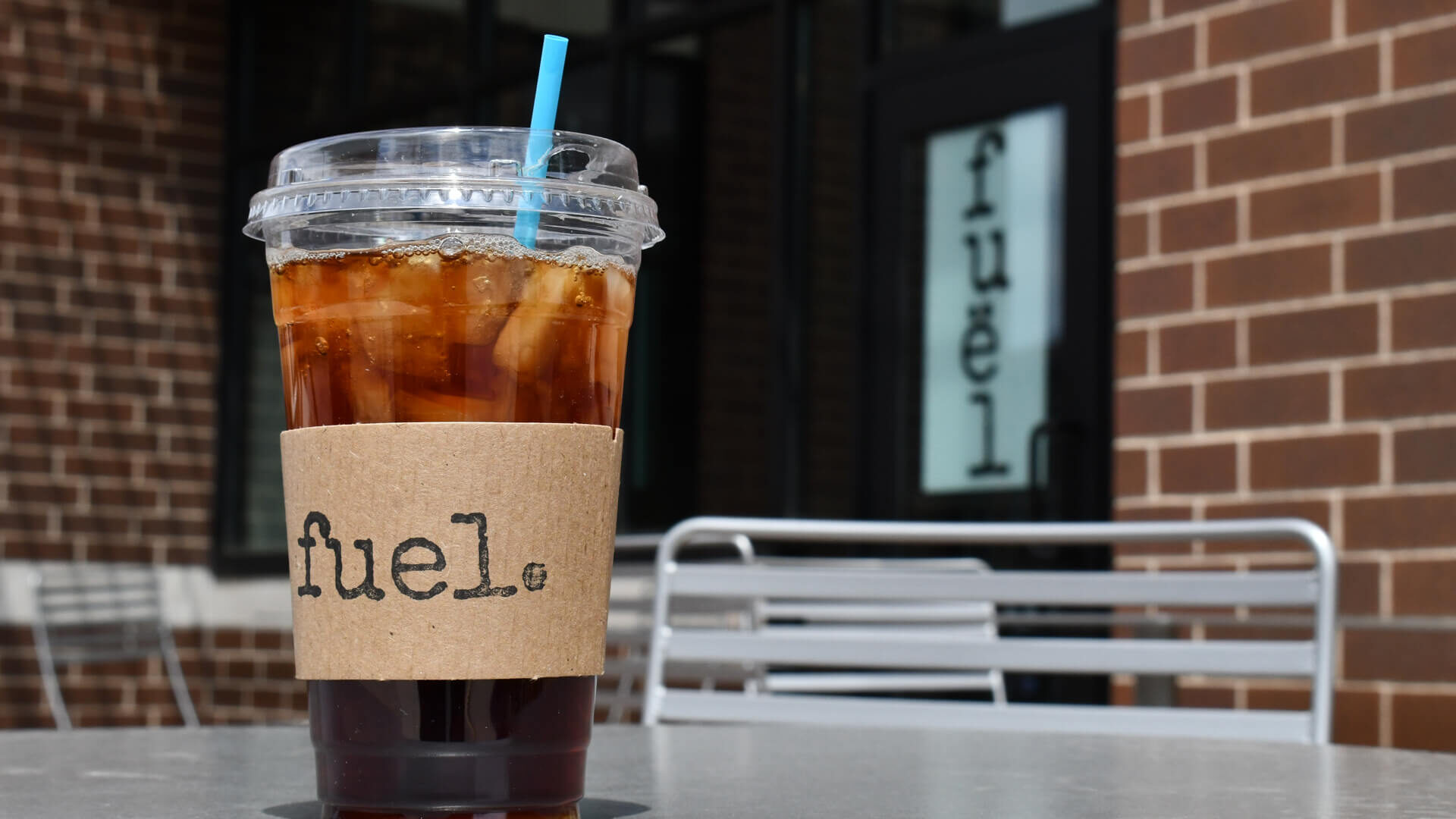 A large ice coffee with a blue straw sits on a cafe table outside of the Fuel coffee shop.