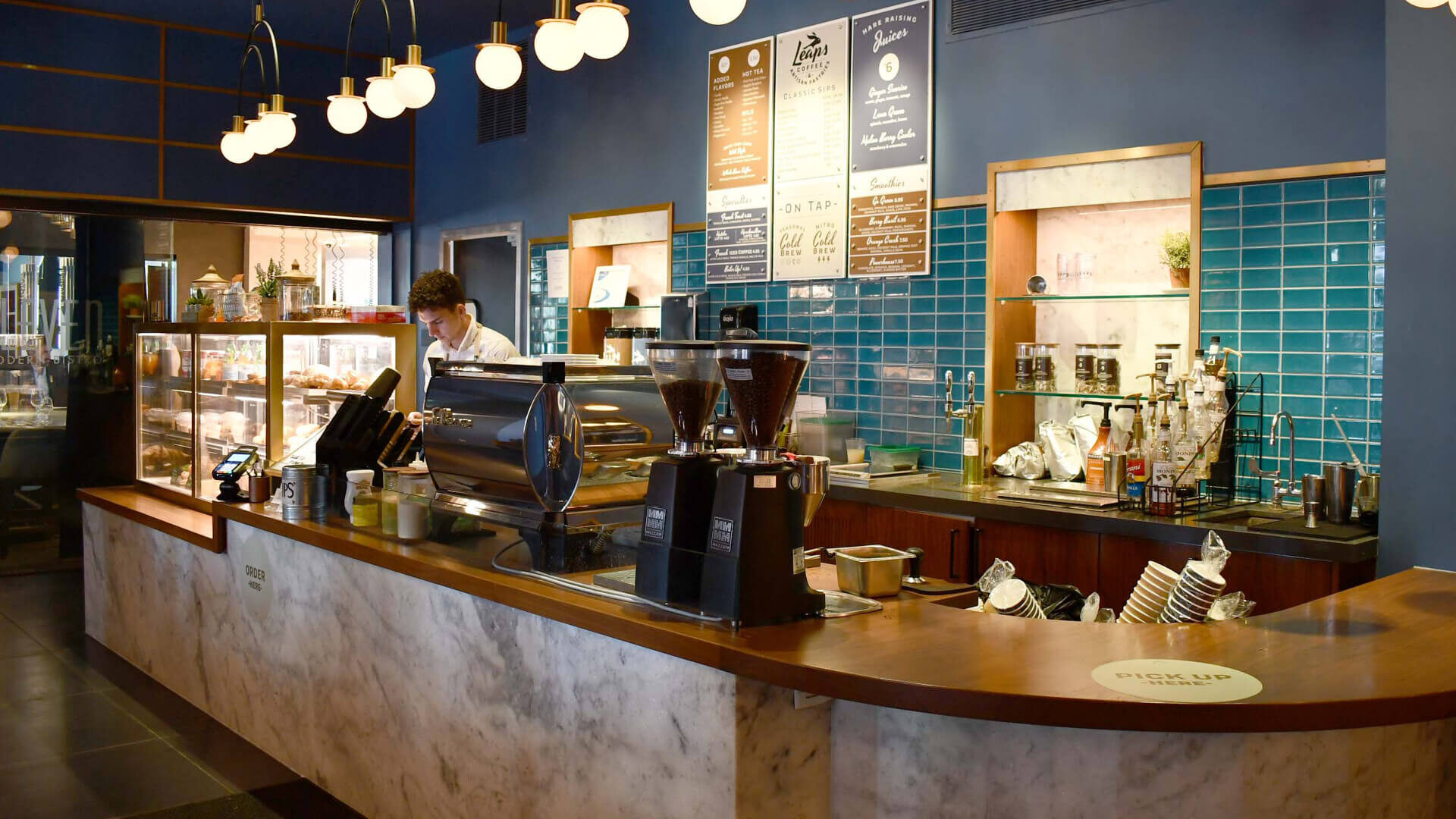 A photo of a student working in the Leaps Coffee bar. The colors of the shop are navy blue, with wood and marble counters and finished with gold lighting fixtures.