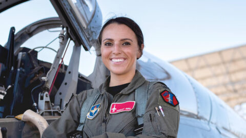 Ashley Bird stands in front of a plane in a green flight suit decorated with patches.