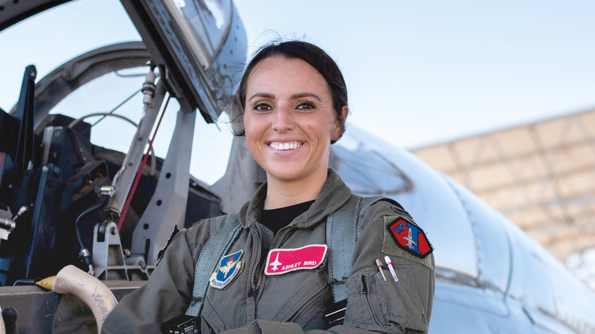 Ashley Bird stands in front of a plane in a green flight suit decorated with patches.