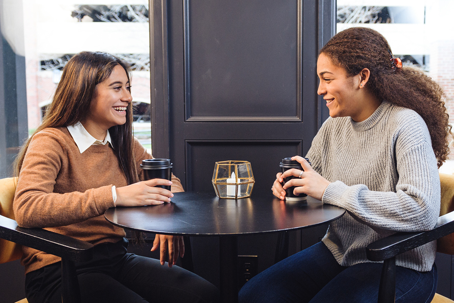 Diane Santos sits across from Kennedy Outlaw at a table. They are both holding coffee cups and Diane is smiling.