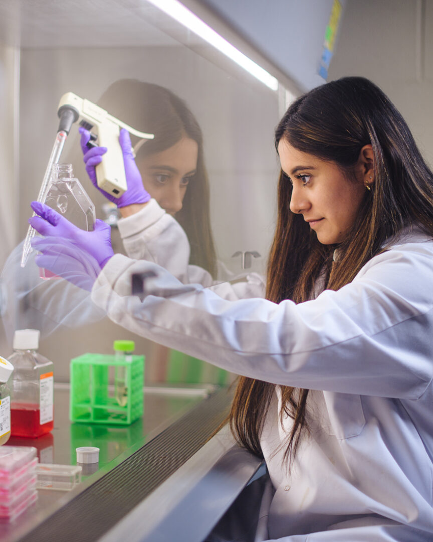 Diane Santos is wearing a white lab coat and holding a piece of science equipment with her right hand and a clear container with her left hand.