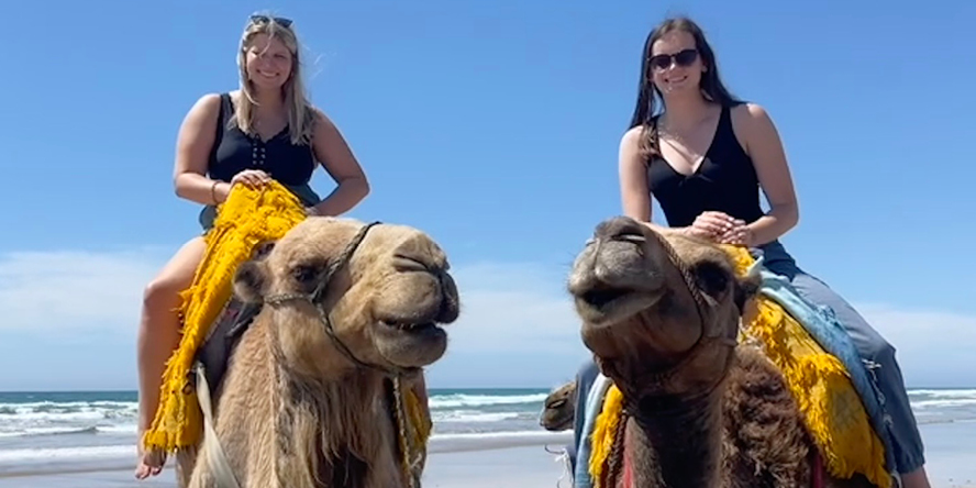 Alexia and her friend are each sitting on their own camel; both camels appear to be smiling