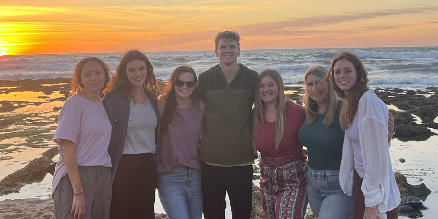 Alexia and six friends pose in front of a sandy beach at sunset