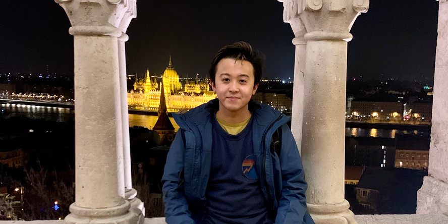 Nick rests against stone arch structure with the Budapest skyline in the background.