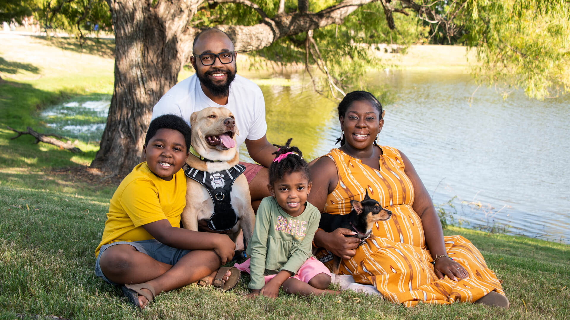 Jacque sits with her family and two dogs near a small lake.