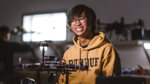 Hudson Tsang smiles at the camera. A small drone sits on the table in front of him.