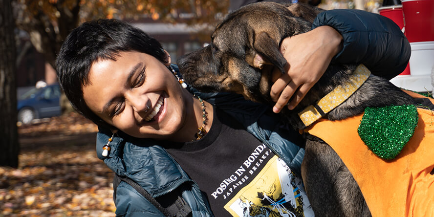 A dog sniffs a student’s neck, who is embracing the dog and laughing