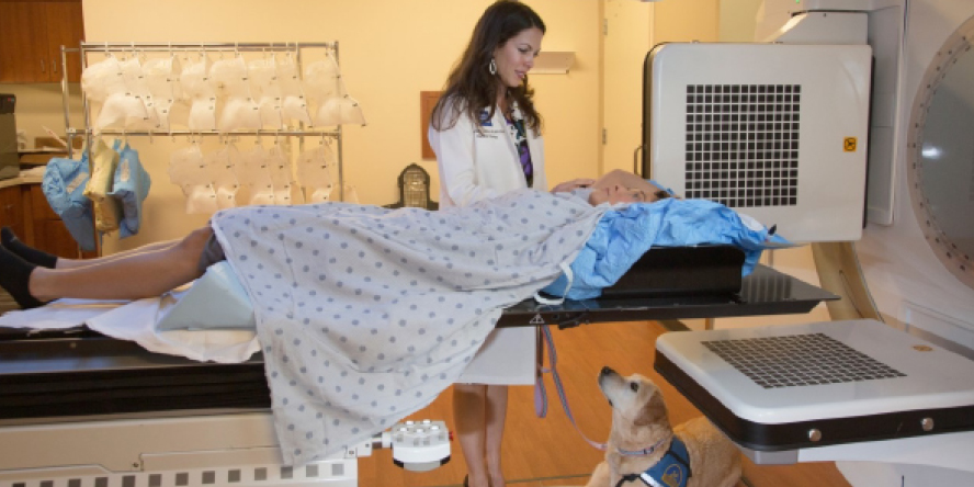 Dr. Michelle Ludwig assists a patient underneath a linear accelerator used in radiation therapy. Ludwig’s service dog, Pam sits on the floor.