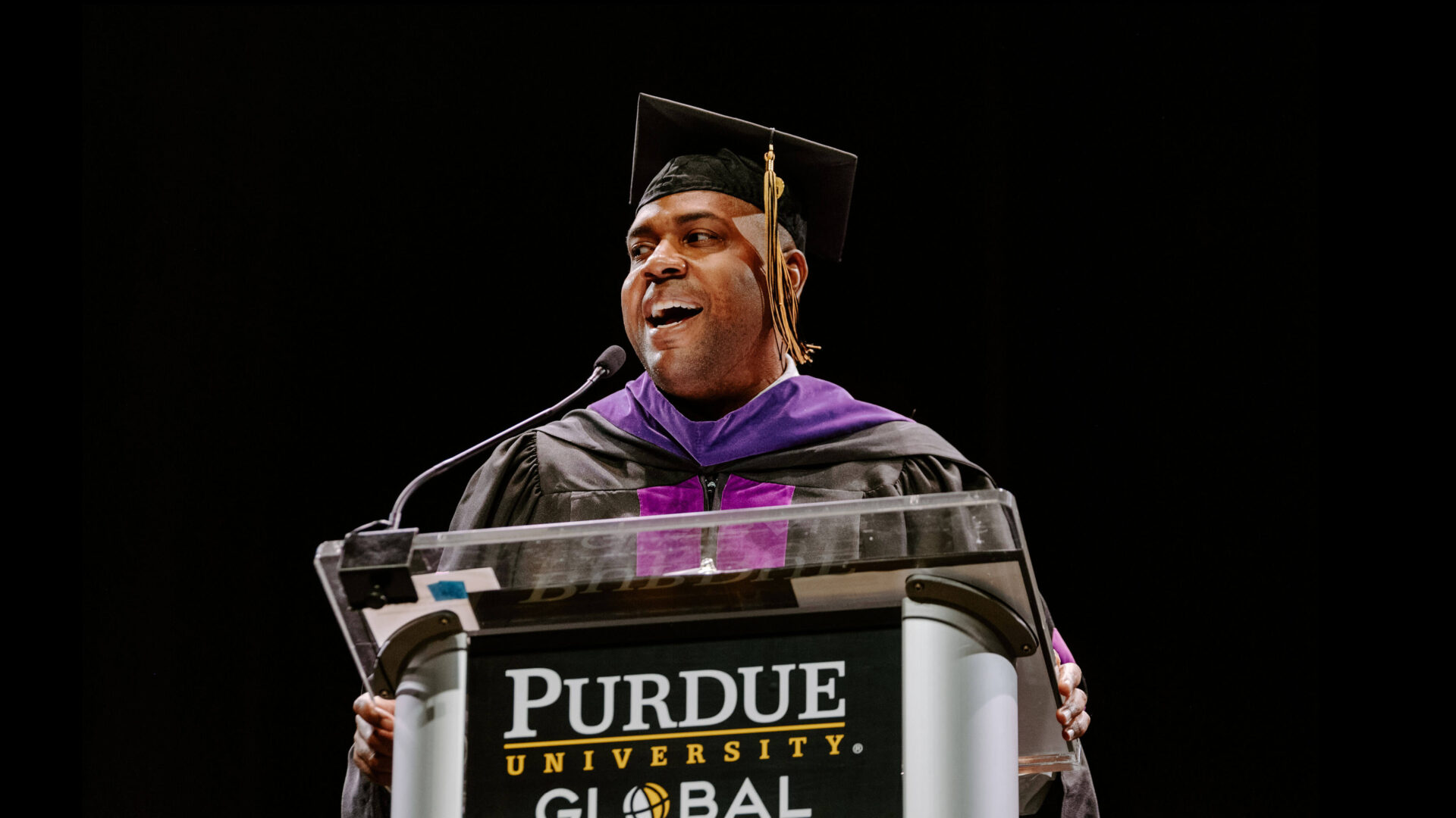 Dolan Williams stands at a Purdue Global lectern with commencement gown and regalia.