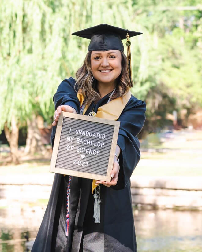 Finch wears her cap and gown and smiles in front of a weeping willow tree, holding a letterboard stating she graduated with her Bachelor of Science, 2023.