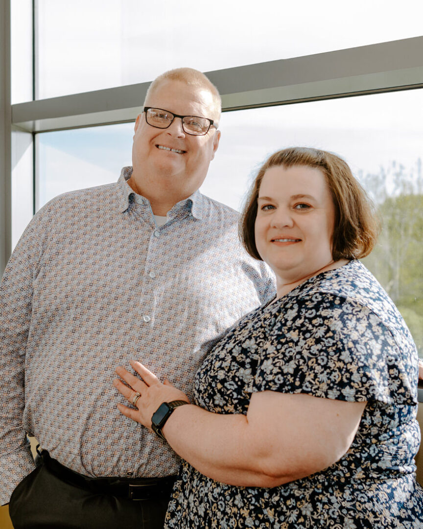 Joe and Donna are smiling; he has his arm around her and her hand rests on his chest, a bright and sunny window behind them.