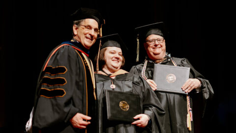 Donna and Joe are crossing the stage together and pause for a photo with Frank Dooley, holding their diplomas.