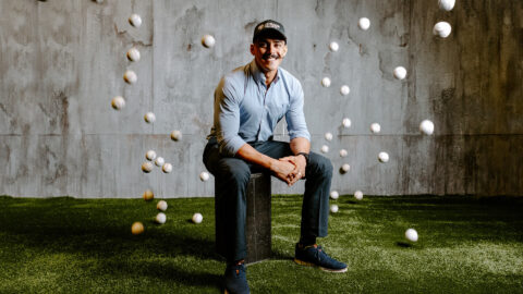 Montes poses casually, hands folded and smiling, wearing a Purdue Global hat, surrounded by flying baseballs.