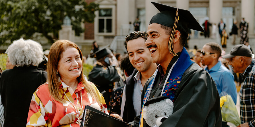 Juan is wearing his cap and gown, holding his diploma and a gray teddy bear wearing a cap, and his mom and brother look at him, smiling.