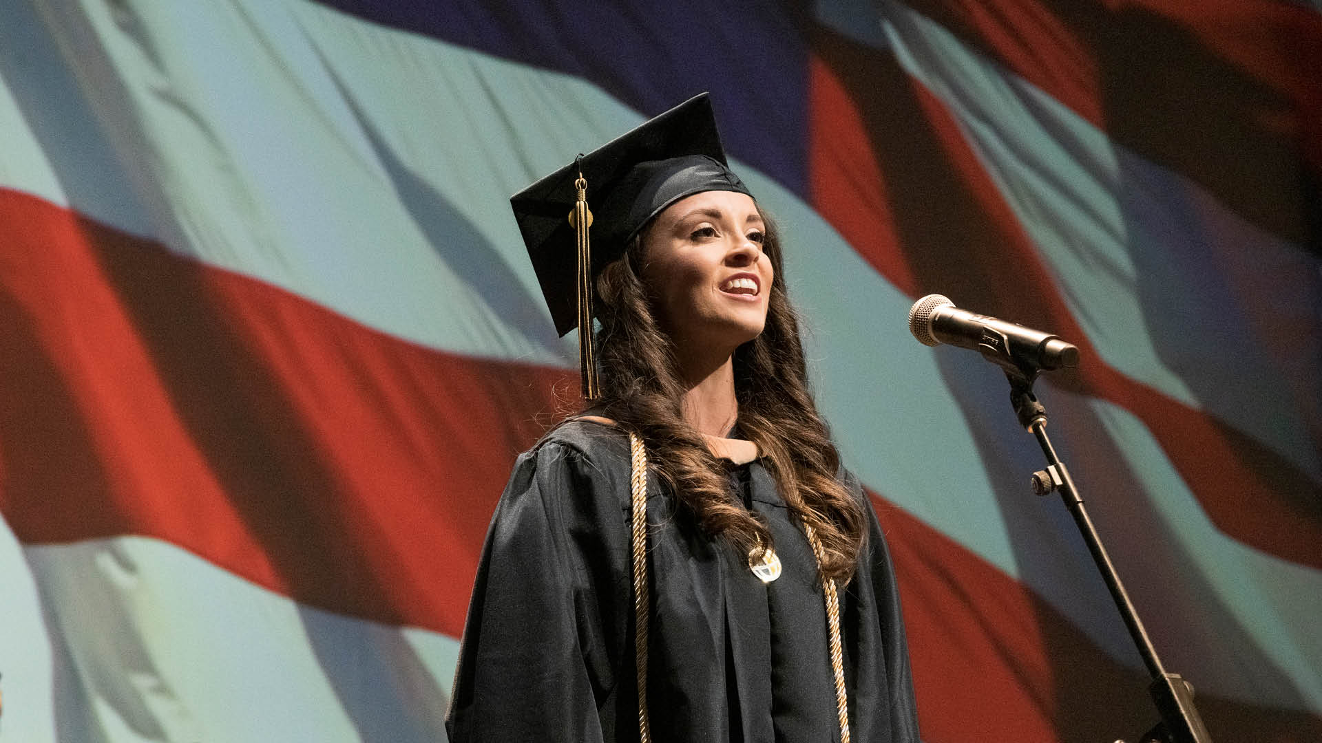 Bruso sings in her cap and gown, with the image of the American flag behind her