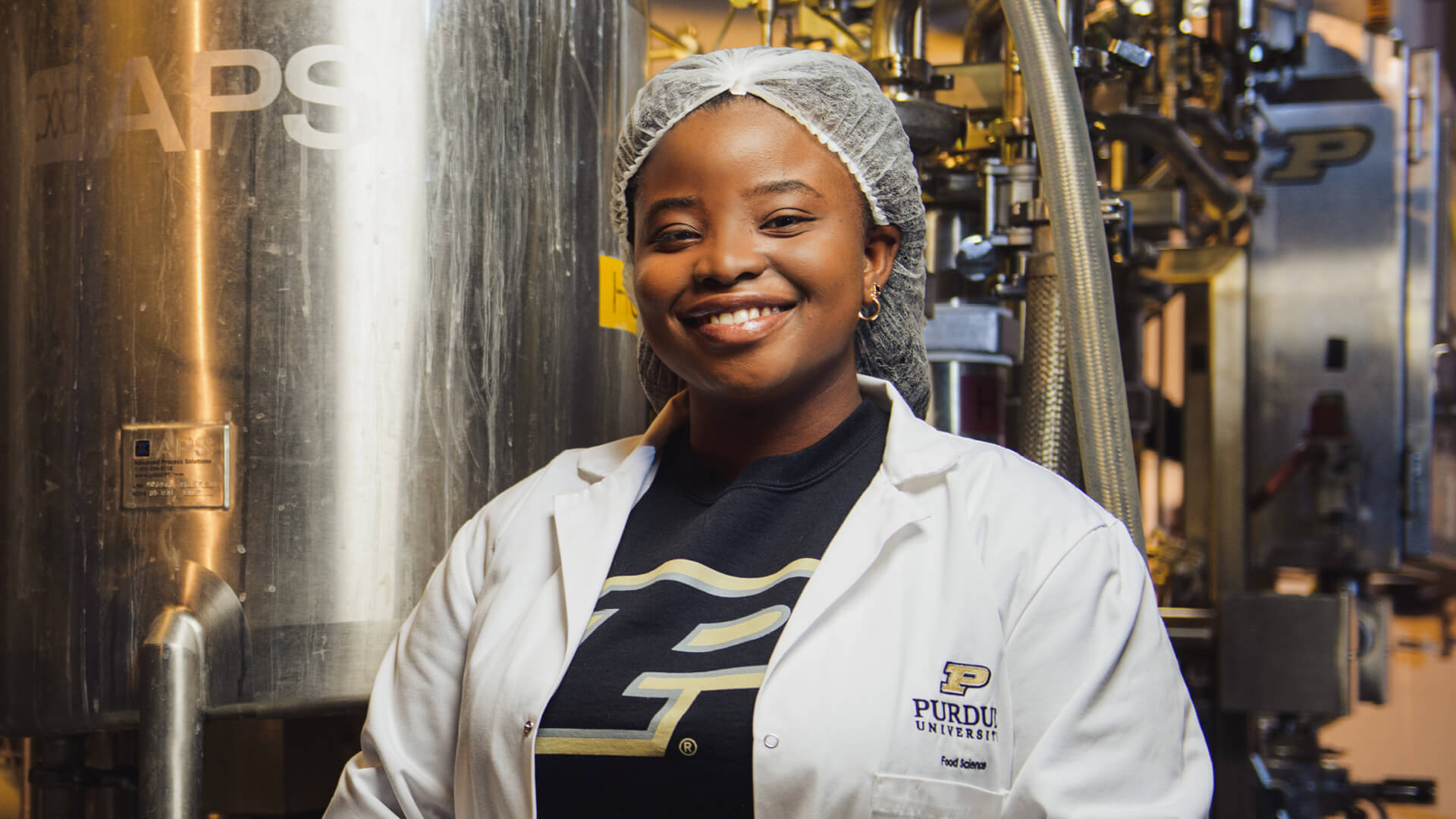 Doriane Sossou smiles at the camera, standing in a lab with metal machinery behind her.