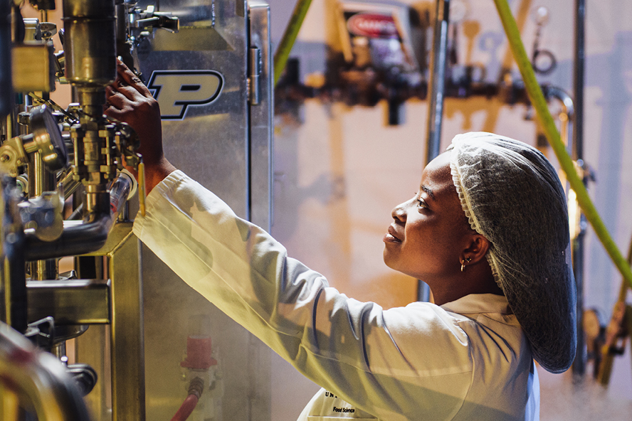 Doriane Sossou, wearing a lab coat and hair net, checks on machinery in a lab.