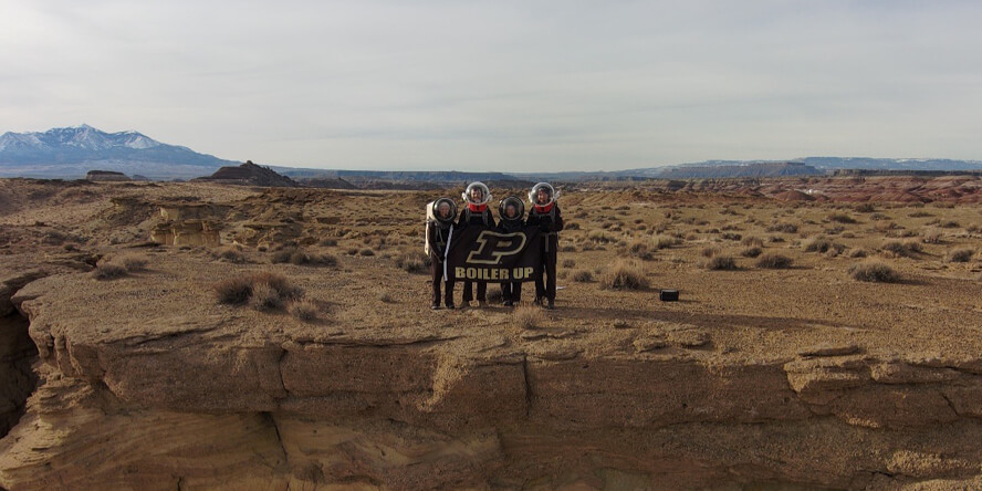 Four members of Crew 272 hold a Purdue flag on top of a cliff.