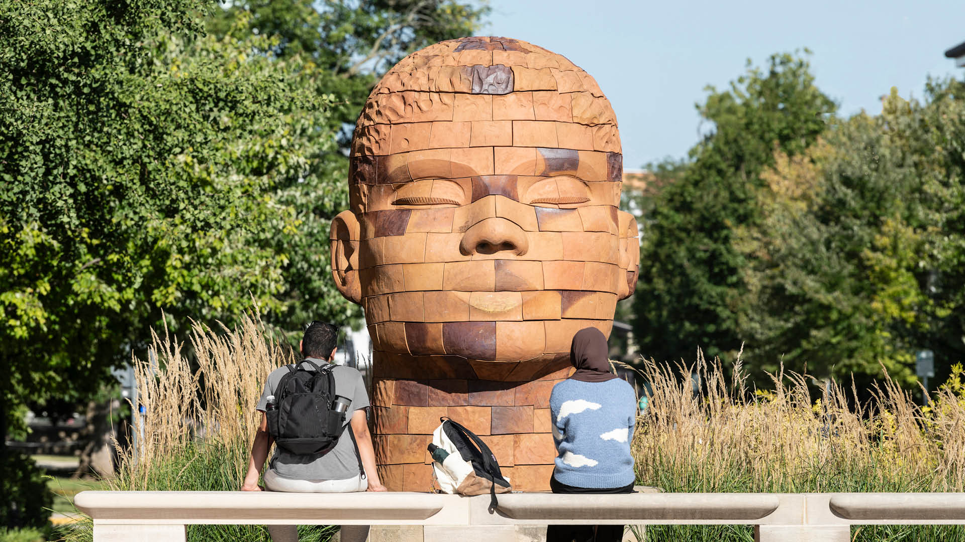 Two students sit in front of a large sculpture of a head made of bricks.