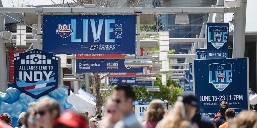 People walking beneath a series of blue USA Swimming LIVE 2024 banners with Purdue signature logo on them.