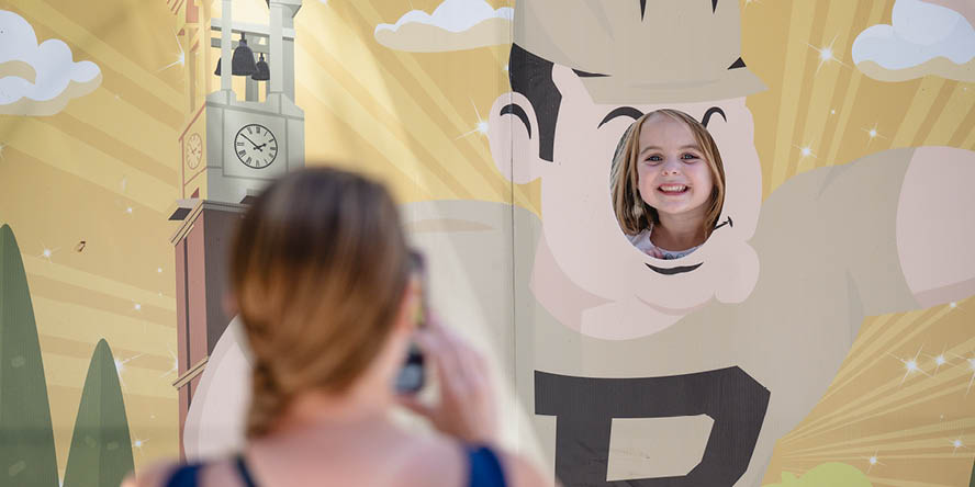 Child with smiling face peering through Purdue Pete cutout, woman in foreground taking a photo with her phone.