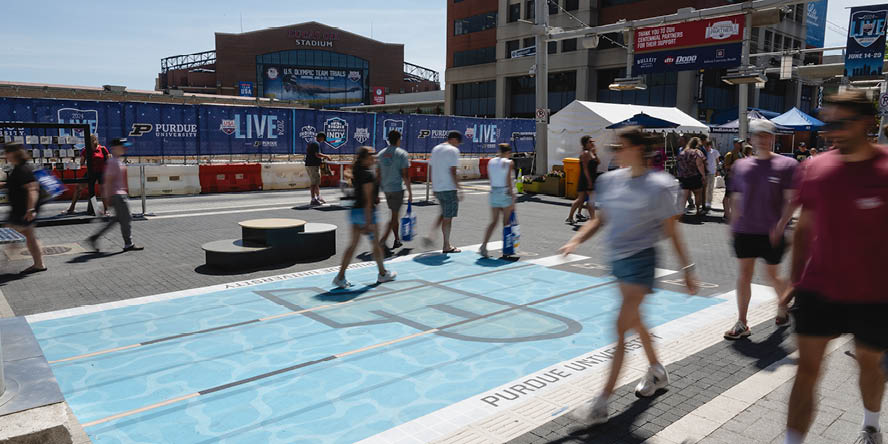 Crowd of people walking in Indianapolis near Purdue-branded swimming pool sidewalk graphic, Lucas Oil Stadium in background.