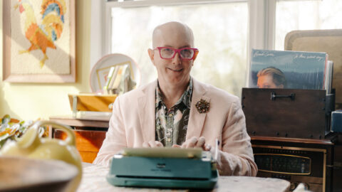 Josef sits at his typewriter in a sunny room, wearing a light linen blazer and a green patterned shirt.