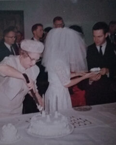 A woman cuts a wedding cake in the foreground. In the background a bride and groom handle cake slices.