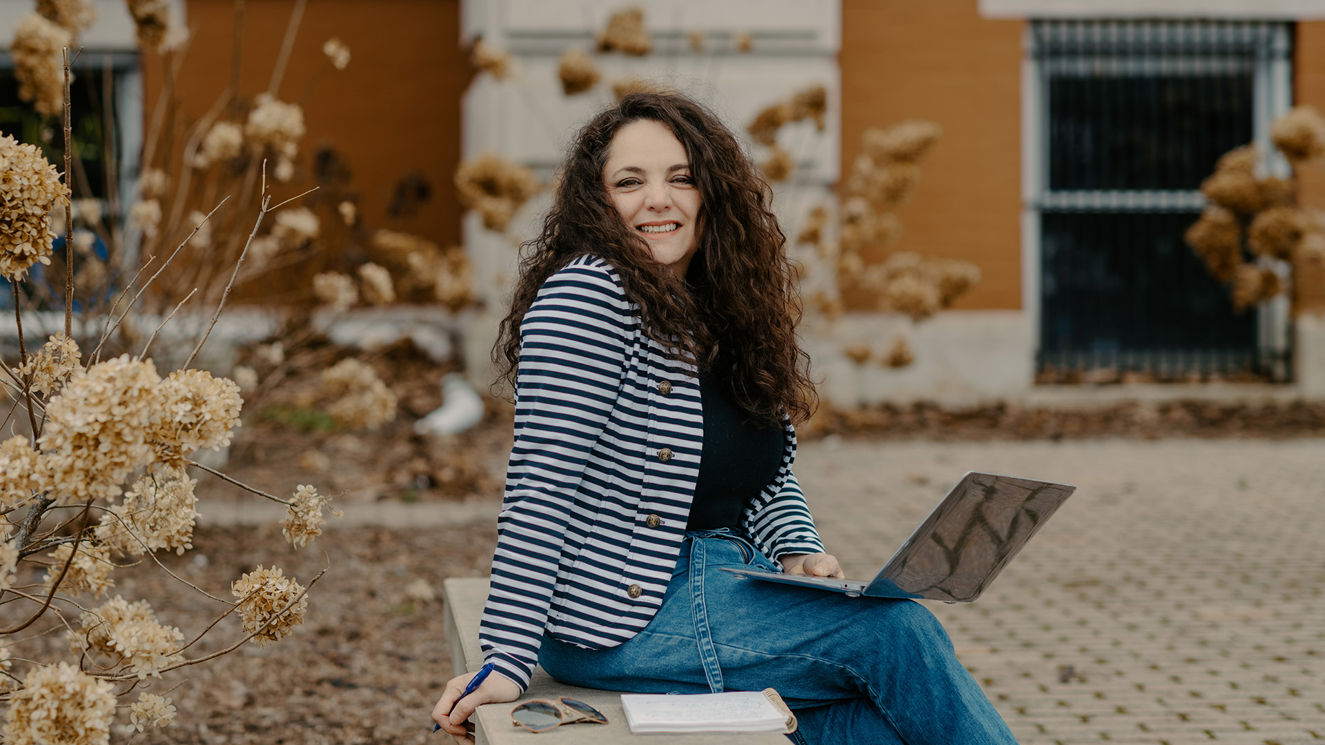 Alejandra Palma sits on a concrete park bench, wearing a navy and white striped jacket, holding an open laptop.