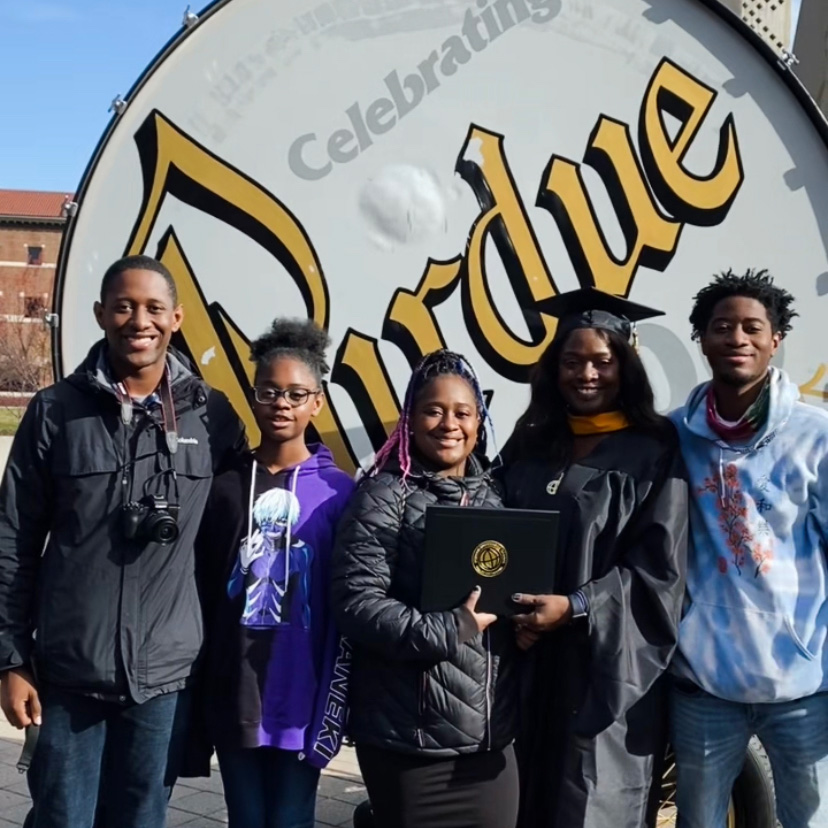 Desiré Hunter poses with her kids in front of Purdue’s Big Bass Drum, wearing her cap, gown and graduation hood, holding her diploma
