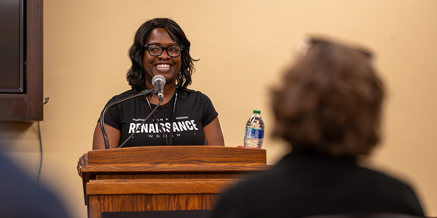 Desiré Hunter stands at a podium, smiling and answering a question from the audience, wearing a Purdue Women’s Conference T-shirt.