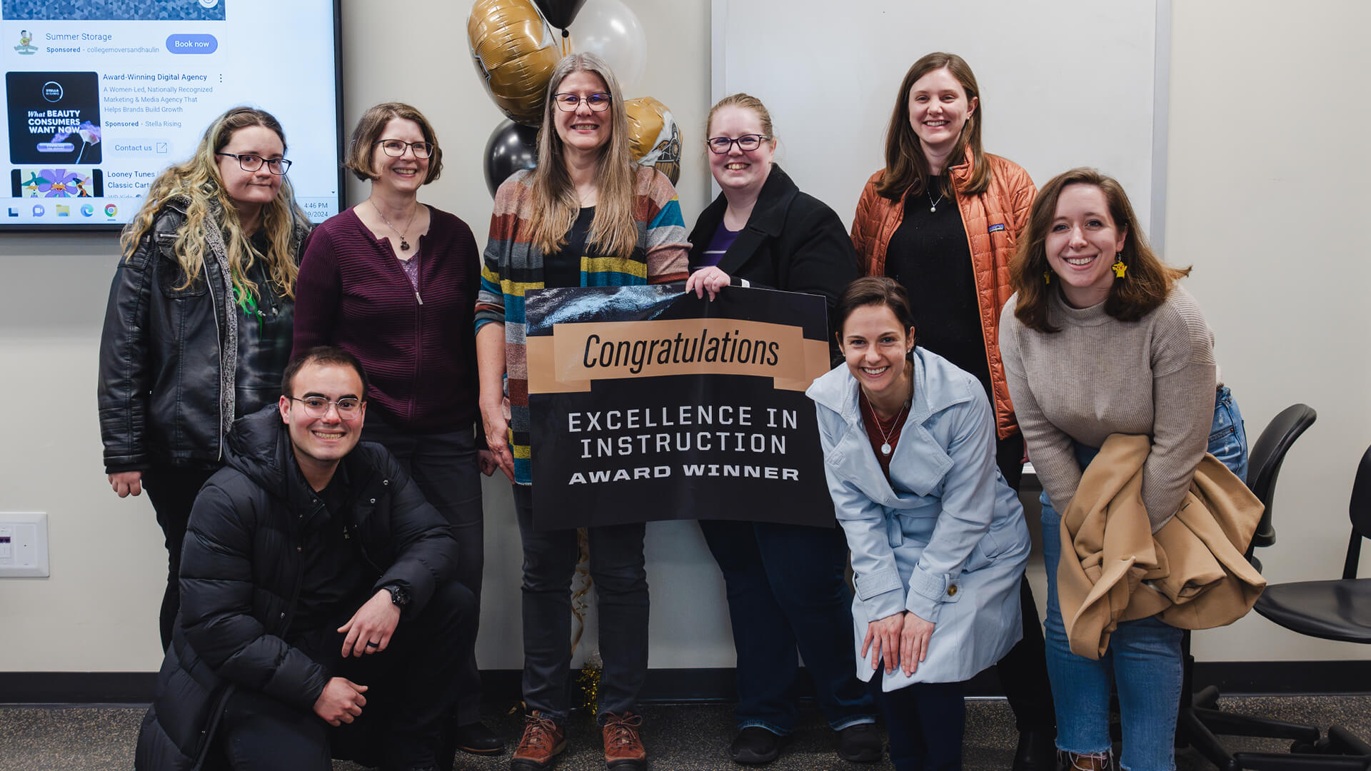 Haynes is wearing a striped cardigan, holding a sign that says, “Congratulations Excellence in Instruction Award Winner,” surrounded by a group of seven people — friends and family. Black, gold and white balloons are visible behind them.