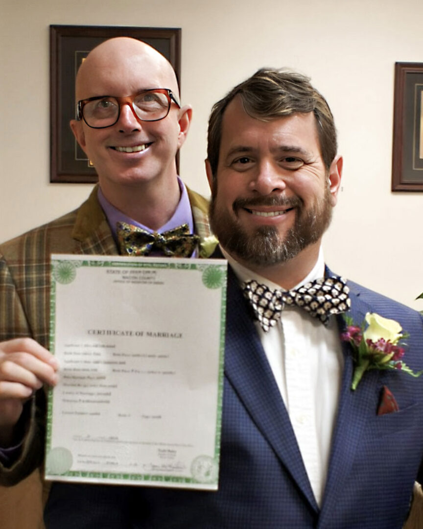 Vice and his husband, both wearing blazers and bowties, smile at the camera and Vice holds up their marriage certificate. 