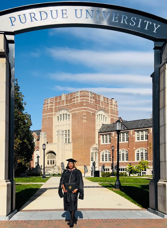 Victoria Durnell standing beneath the Purdue University arch wearing her graduation cap and gown. 