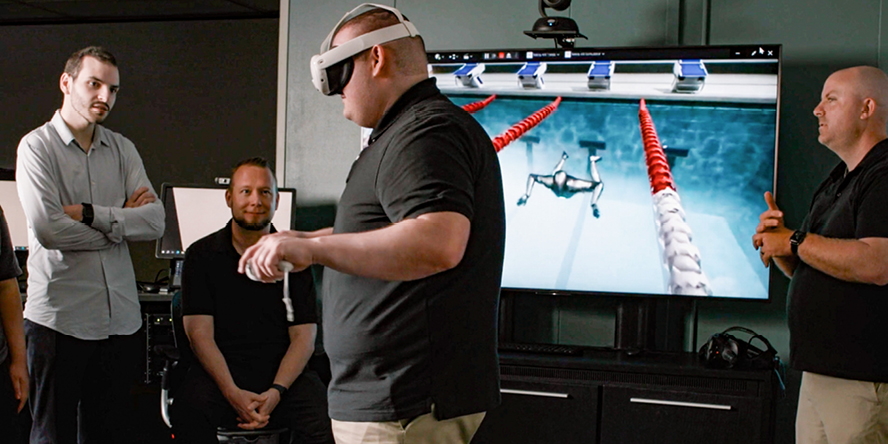 Four men in front of a large screen displaying an animation of a swimmer. One man wearing a VR headset.