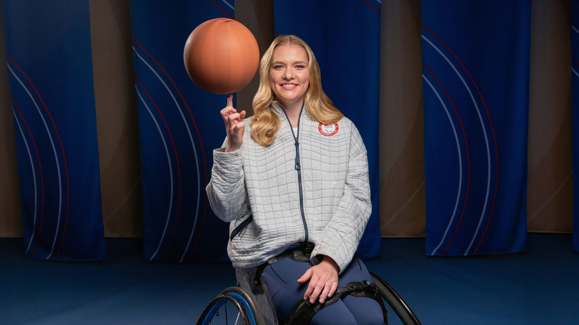 Rose Hollermann, wearing a Team USA jacket, is spinning a basketball on one finger and smiling.