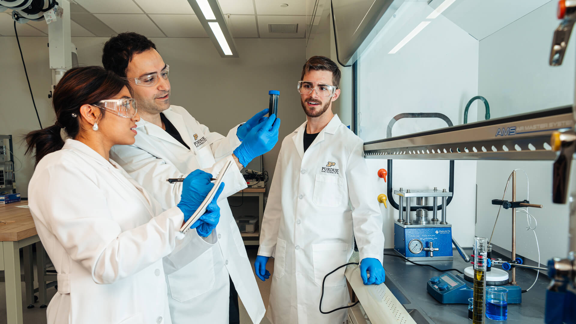 A professor holds up a test tube in a lab while one student takes notes and another looks on.