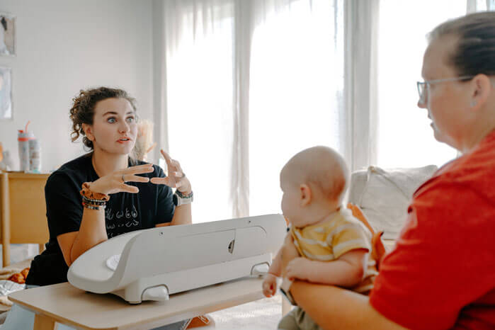 Sienna is discussing something with a client, who sits on a white couch holding her infant. 