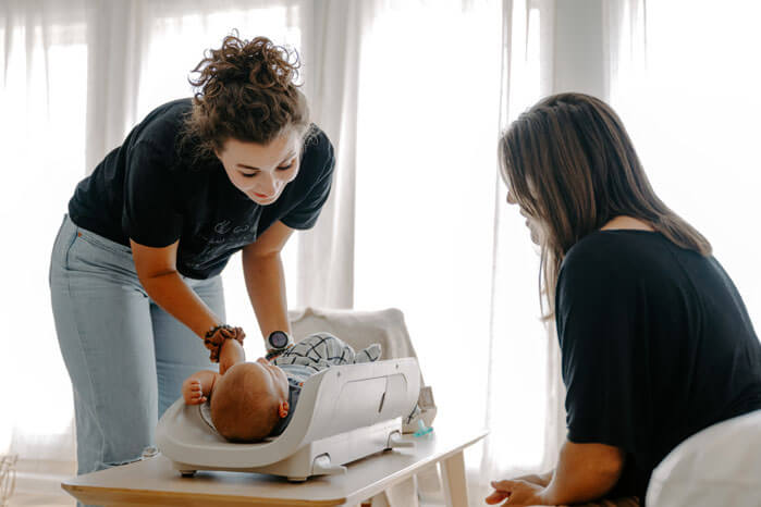 Sienna stands to weigh a baby while the mother’s back is visible, seated on a couch. 