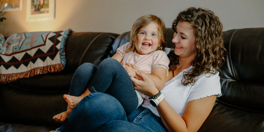 Sienna is sitting on a black leather couch with a giggling toddler on her lap. 