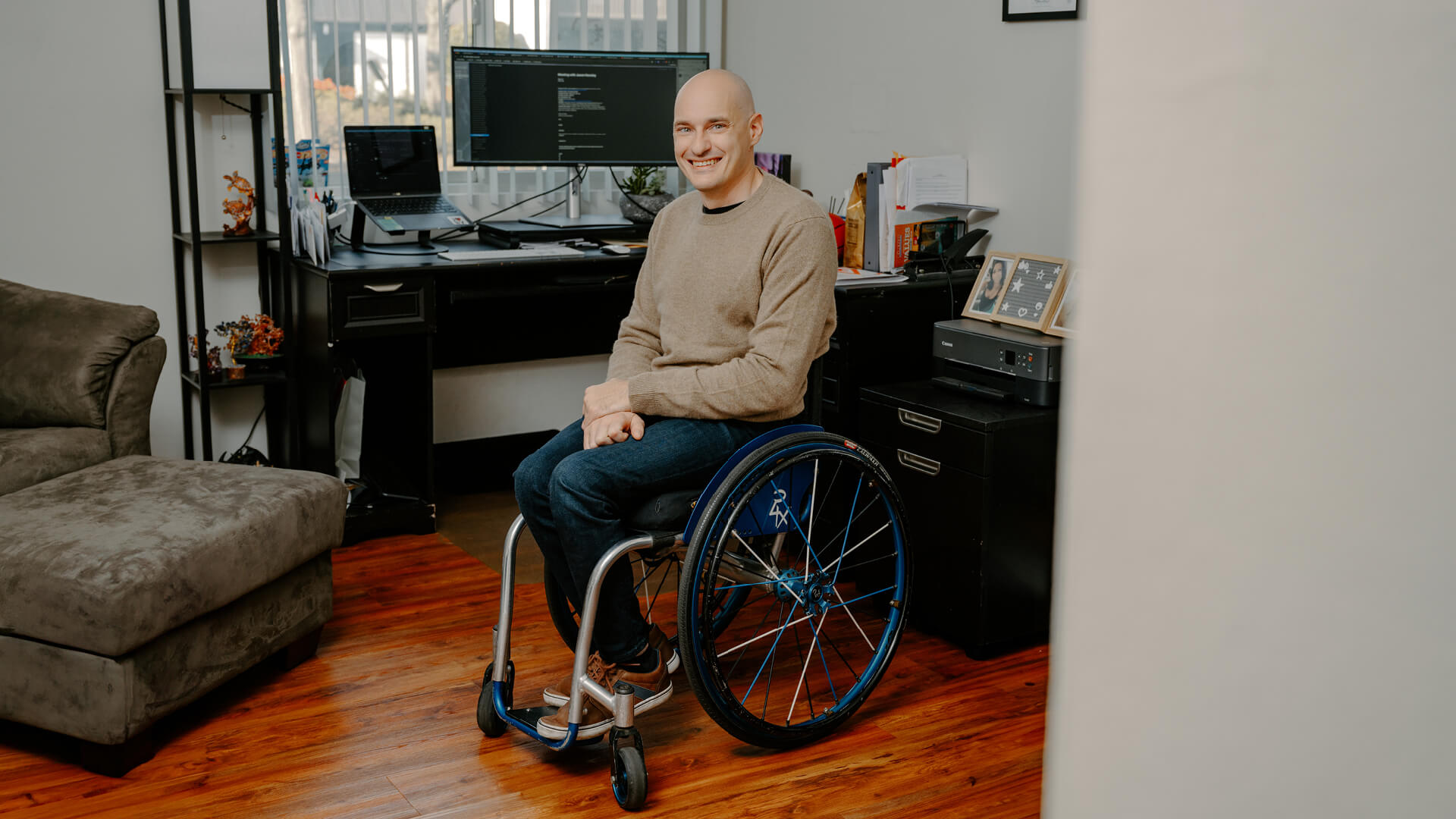 Kevin is seated in his wheelchair, smiling, with his desk and computer behind him. 