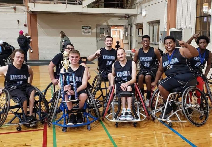 Kevin is one of eight men pictured in their wheelchairs, wearing blue jerseys that read “LWSRA,” holding a trophy on a basketball court. 