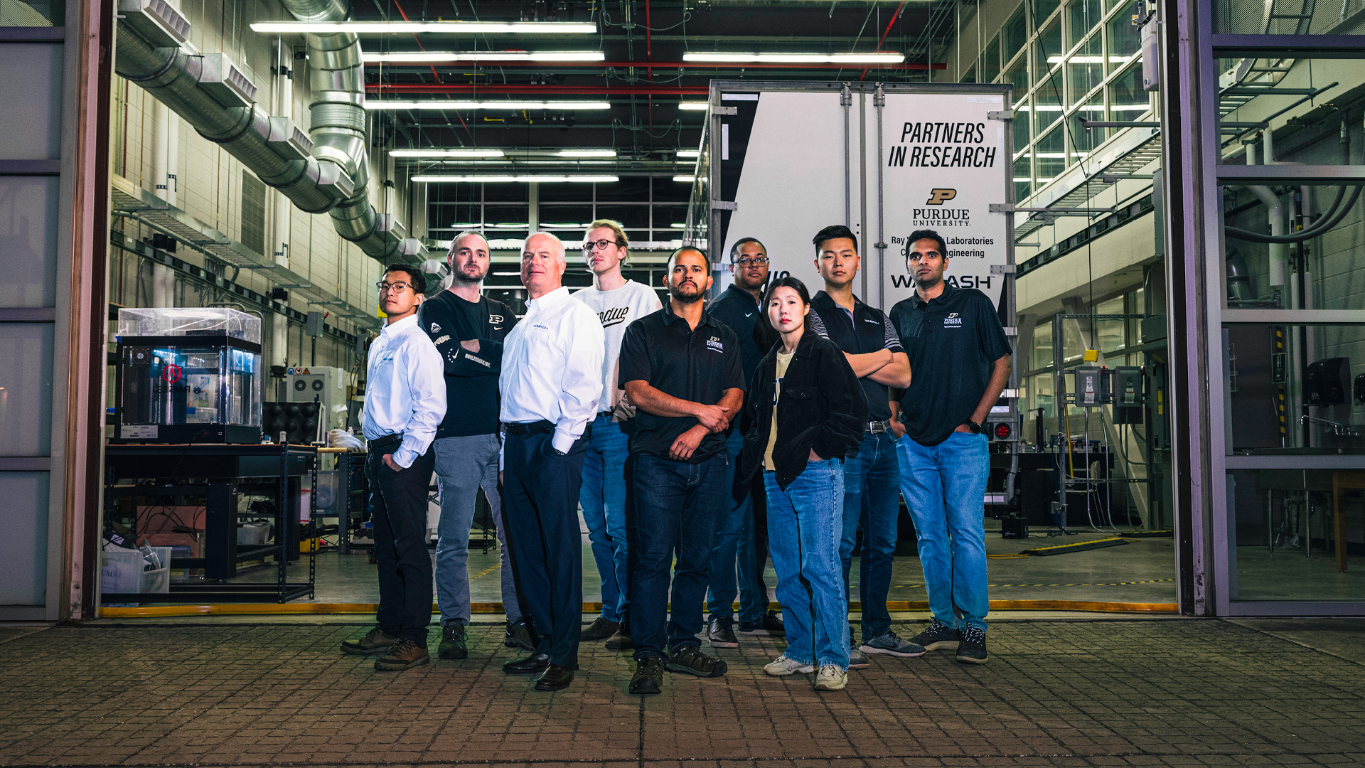 Eight men and one woman standing in the open door of a large lab with a semi-trailer in the background.