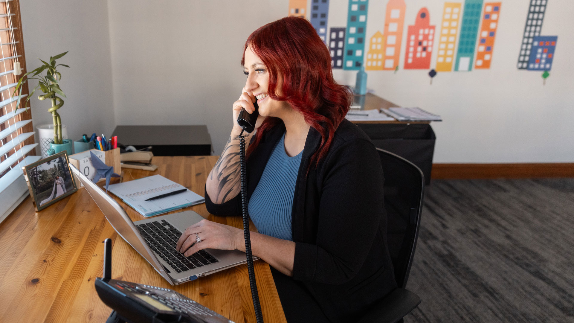 Melton is at the same desk, viewed from the side, with a desk phone at her ear and looking at her laptop.