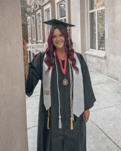 Tara is wearing her cap and gown, posing on campus. She’s leaning against a limestone pillar, a building with windows behind her.