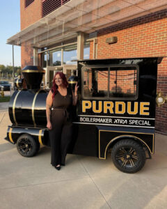 Tara smiles and stands next to a small, black engine at sunset.