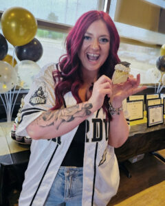 Tara is wearing a white button-front Purdue baseball jersey and is holding up a celebratory cupcake.